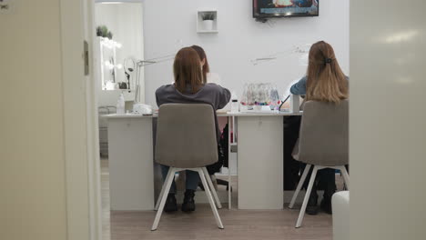 two female customers seated facing nail technicians inside bright nail salon with overhead lamps, manicure tools, sample palettes on desk, visible television on wall, and modern white decoration