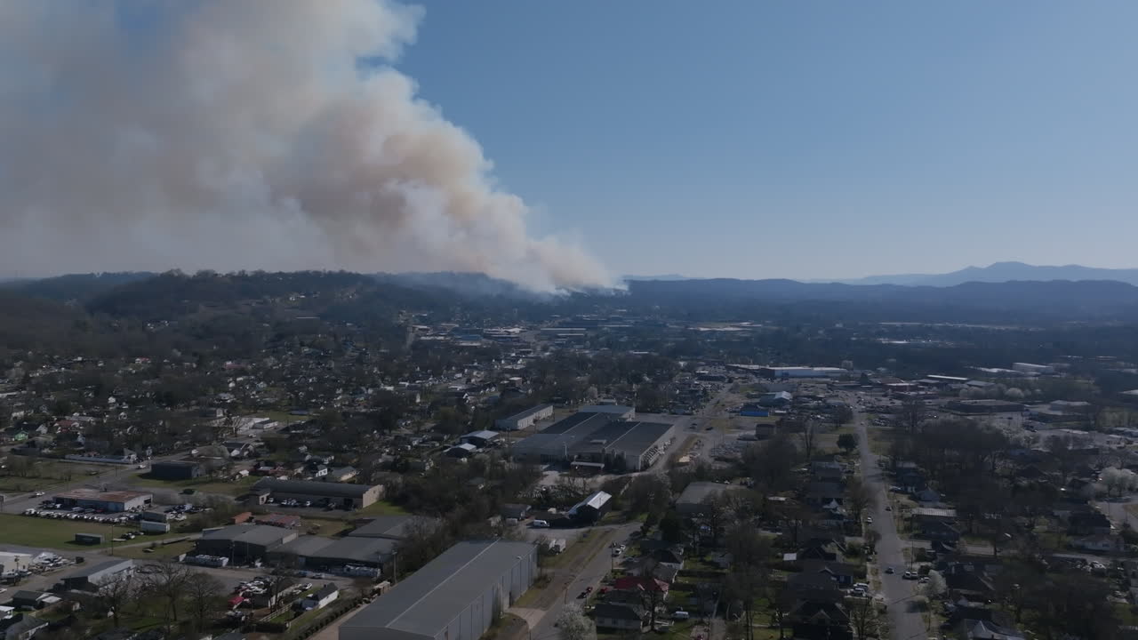 Aerial drone footage flying over an industrial area of a city with a large wildfire going on in the background.