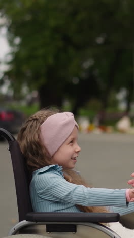 cheerful girl with arthritis enjoys playing game with mom after walk in park. redhead woman has fun with little daughter in wheelchair closeup