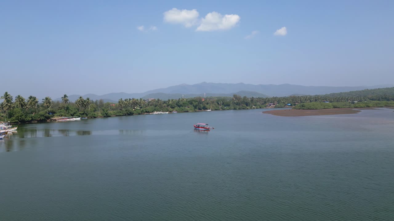 Sunny day at Rajbag Beach, Goa boats gently parked along the serene Talpona River. Lush green trees line the shore, with distant mountains creating a stunning tropical backdrop, Drone