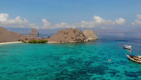 Vuelo-Panorámico-Desde-Barcos-Turísticos-De-Madera-Sobre-Las-Aguas-Turquesas-De-La-Playa-Rosada-Hacia-Las-Empinadas-Colinas-De-La-Isla-Padar-En-El-Parque-Nacional-De-Komodo,-Indonesia