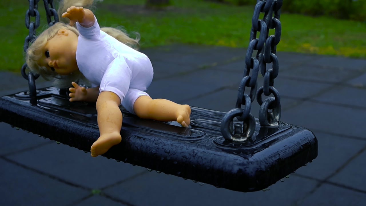 Bambola triste e sola con capelli biondi sdraiata su un'altalena bagnata in un parco giochi durante una giornata di pioggia, creando un'atmosfera malinconica e inquietante con una sensazione di abbandono infantile.