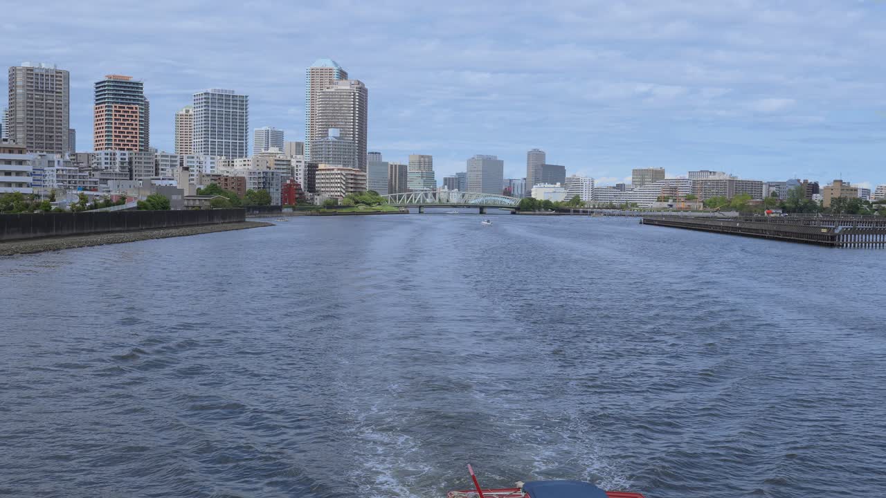 A peaceful shot of a passenger boat traveling down a river with the Tokyo cityscape and modern buildings in the background