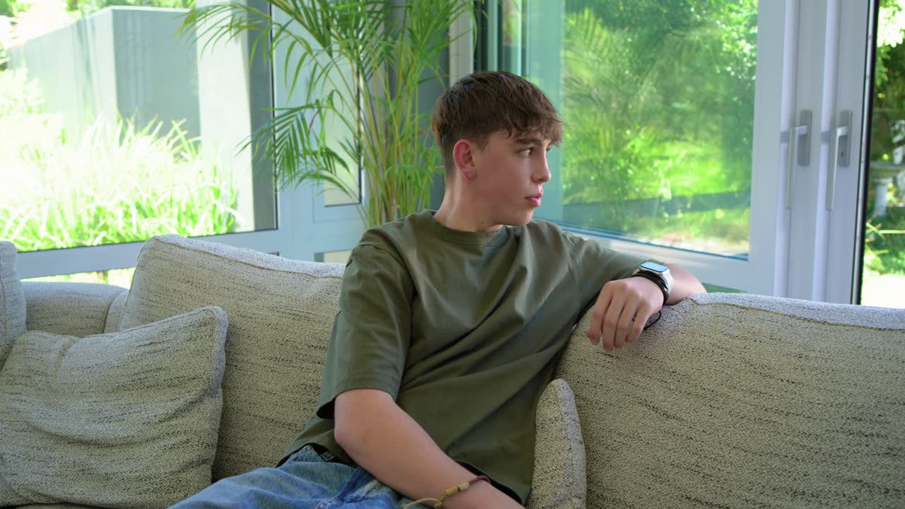 Teenage male noticing camera, turning, sitting on beige sofa by corner window, smiling at camera