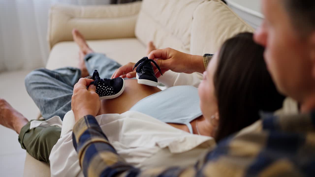 행복한 중년 남자의 어깨를 가로질러 임신 중인 아내를 만지고 임신 중에 집에 있는 어린 아이에게 작은 신발을 선물합니다.