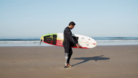 Seitenansicht-Ein-Männlicher-Surfer-Mit-Beinprothese,-Der-Ein-Surfbrett-Unter-Dem-Arm-Hält-Und-Am-Strand-Entlang-Geht