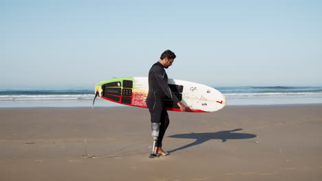 vista lateral de un surfista masculino con una pierna artificial sosteniendo una tabla de surf bajo el brazo y caminando por la playa