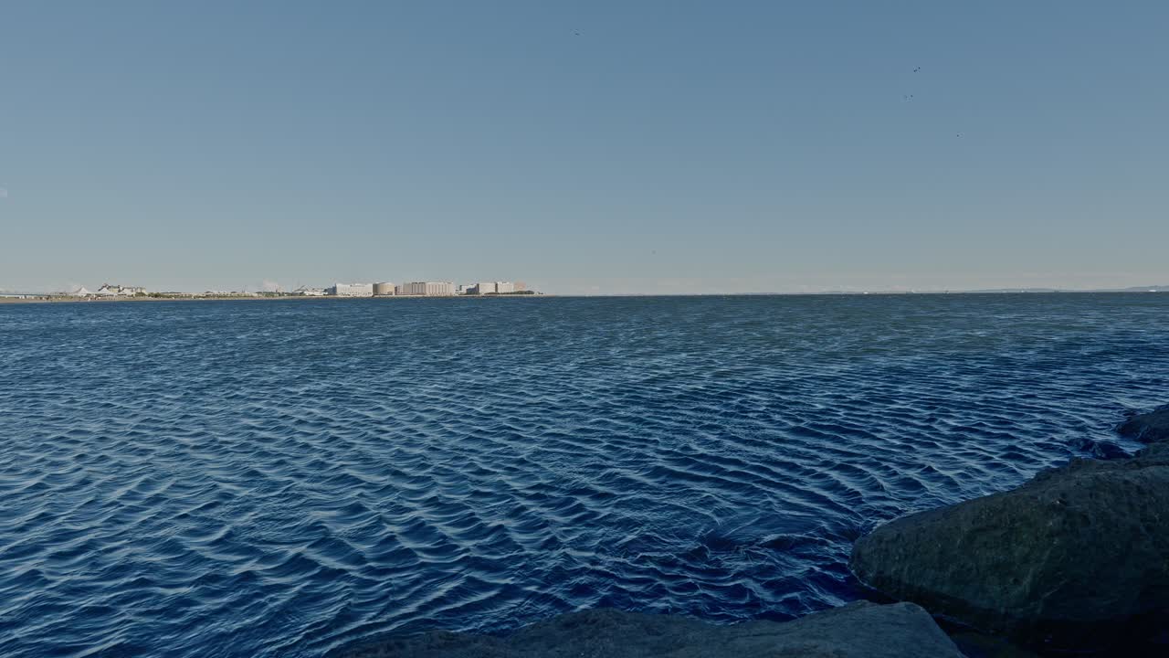 A wide, bright shot of dark blue water with subtle ripples, framed by rocks in the foreground and a line of low-rise buildings on the coast