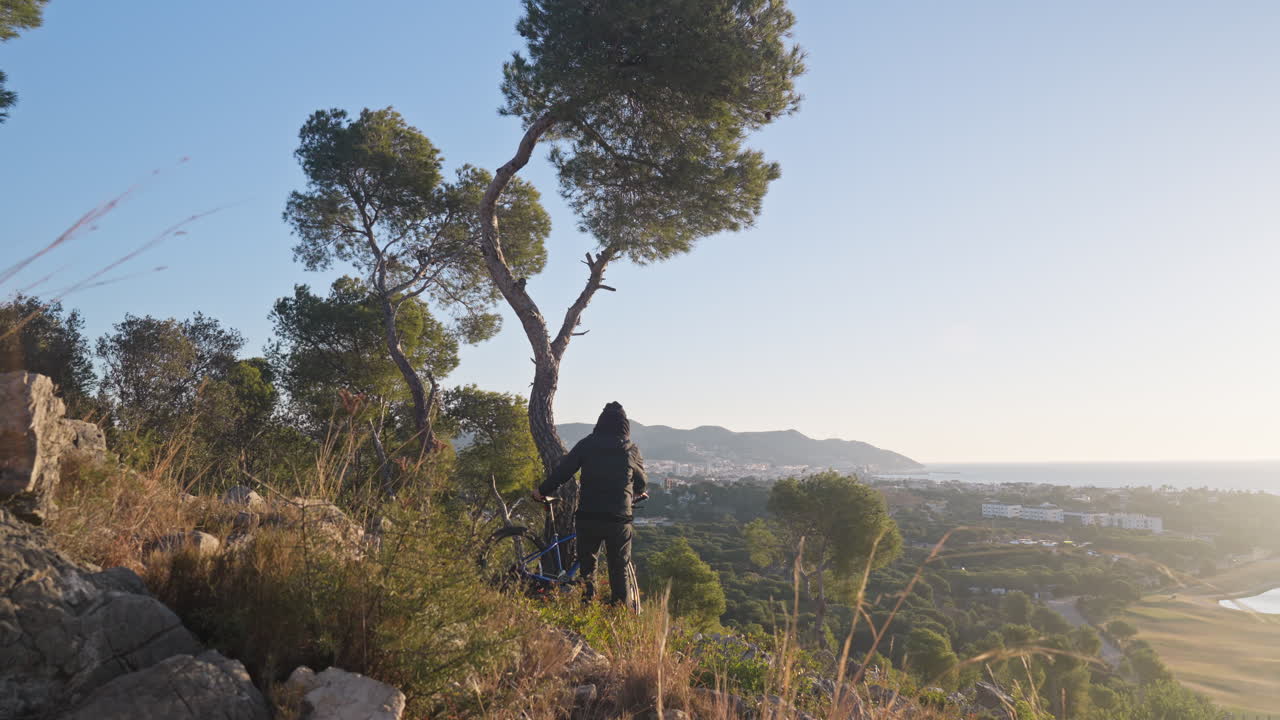 During early morning sunrise with a stunning landscape backdrop, a man walks towards his mountain bike and then departs, pushing bicycle downhill