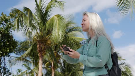 beautiful slim woman with long blonde hair in green shirt standing near palm tree and using smartphone over background the park. girl on the square touching screen and smile