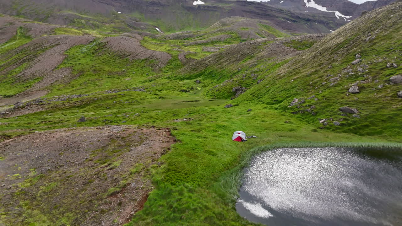 Aerial view of a coastal hillside in Fjallabyggð showing two tents camped beside a small pond, with green mossy terrain, rocky slopes, and the open ocean stretching beyond the winding road
