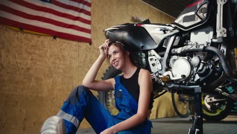 A-tired-girl-auto-mechanic-in-a-black-top-and-blue-overalls-leans-on-a-motorcycle-that-she-jacked-up-and-rests-in-her-workshop-with-a-US-flag-on-the-wall