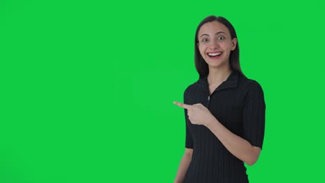 Happy-Indian-female-news-anchor-pointing-at-green-screen-and-showing-thumbs-up