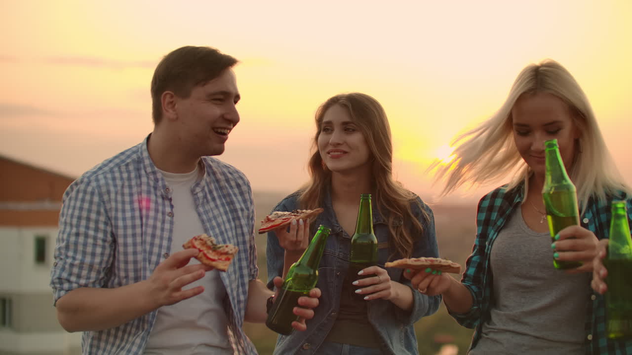 Loving couple drinks beer and eats hot pizza on the roof. Girl gently puts her head on her boyfriend's shoulder. They are smile and enjoy the time at the sunset.