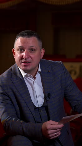 Host announces future theater season. Actor is sitting in empty theater on red velvet chair. Classic theater interior in red colors. Vertical video