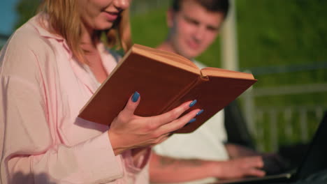 close-up of lady in pink holding book and reading while her friend in background types on laptop and nods in agreement, background includes green hill, outdoor bench, and pole in warm sunlight