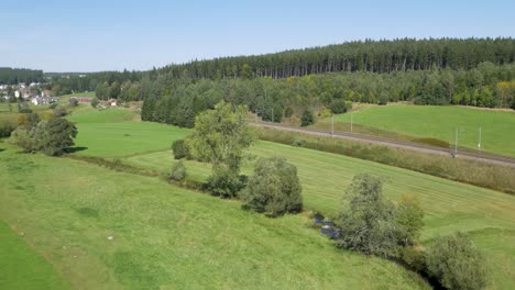 drone shot of a red and white train journeying on a curving track through a lush green valley, dotted with trees and scattered buildings, all under an unblemished blue sky