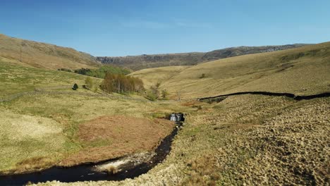 Aerial-shot-close-to-the-ground-of-a-stream-which-feeds-the-Kinder-Reservoir,-Peak-District,-UK-with-the-Kinder-Scout-Peak-and-Waterfall-in-the-distance