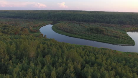 A-drone-flies-low-over-a-pristine-forest-and-calm,-winding-river-valley-at-sunset