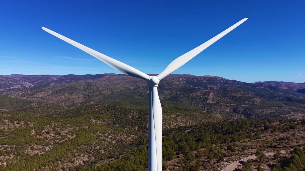 Primo piano di una turbina eolica bianca che gira contro un cielo azzurro limpido sopra montagne spagnole boscose, mostrando l'infrastruttura di energia rinnovabile in un paesaggio naturale