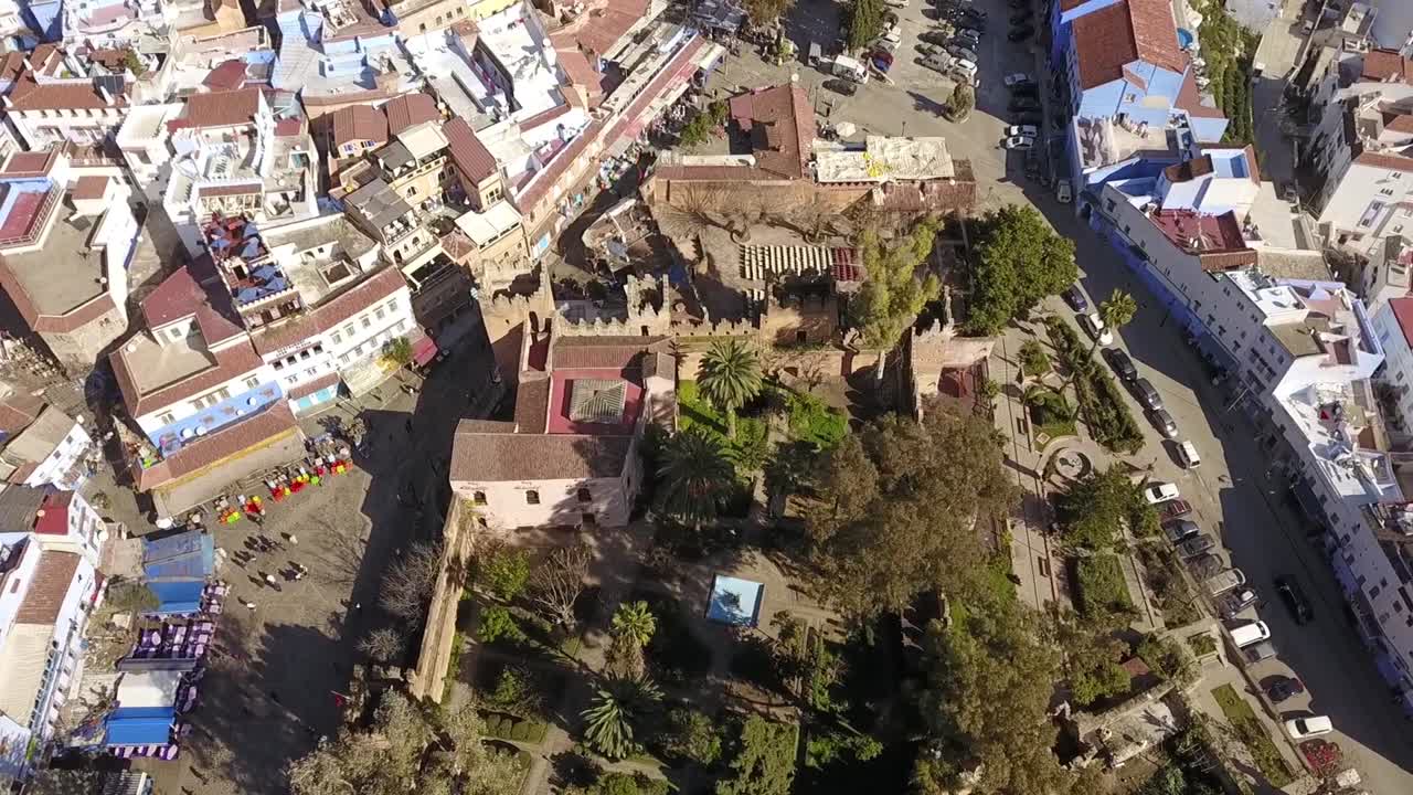 Detaljeret HD luftfoto oppefra af den historiske Kasbah i Chefchaouen med dens røde mure og frodige have. Viser kontrasten mellem fæstningen og medinaens omkringliggende ikoniske blå bygninger