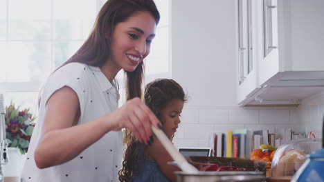 mujer hispana cocinando en la estufa en la cocina con su hija pequeña a su lado, de cerca, vista lateral