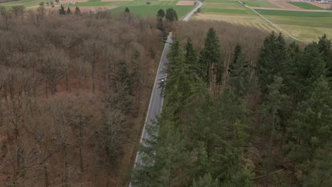 Silver-car-travelling-along-an-empty-country-road-that-leads-through-bare-deciduous-trees-on-the-one-side-and-a-dark-green-coniferous-forest-on-the-other