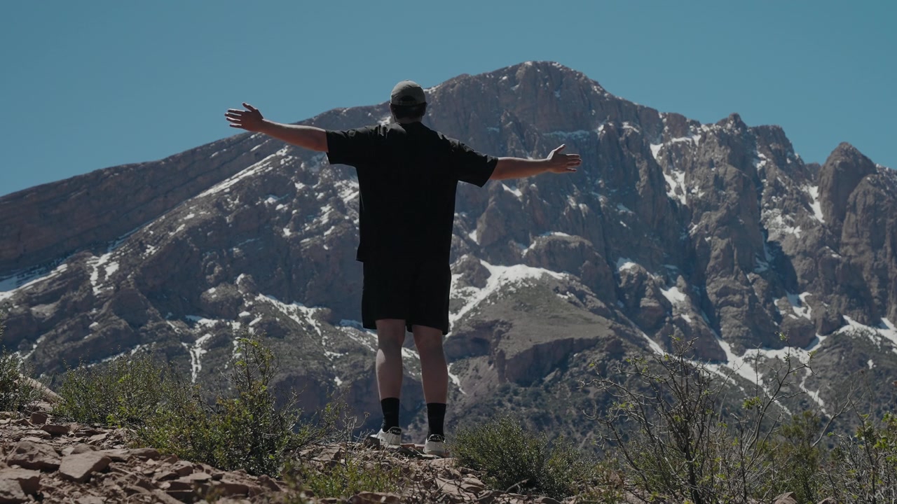 Hombre de pie con los brazos extendidos frente a una escarpada montaña nevada en el Alto Atlas de Marruecos