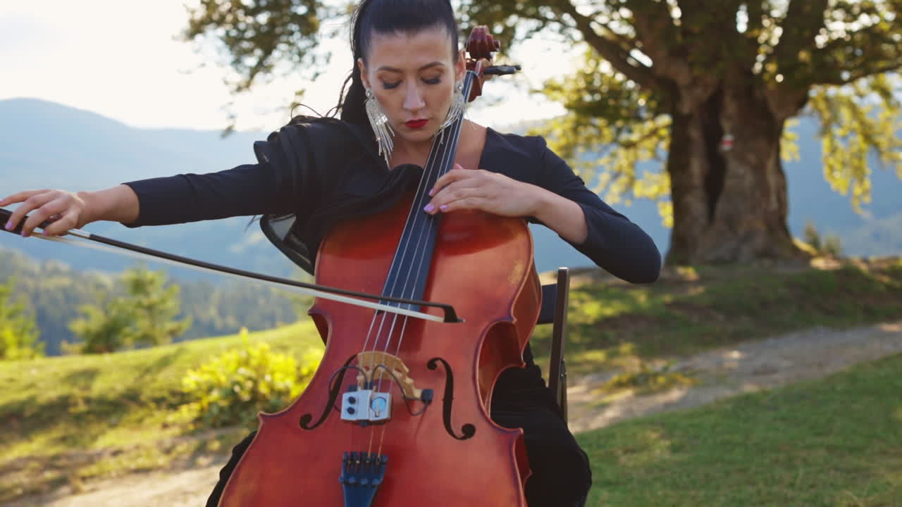 Dark-haired female musician focused on playing cello. Middle-aged woman in black dress plays instrument in the nature backdrop.
