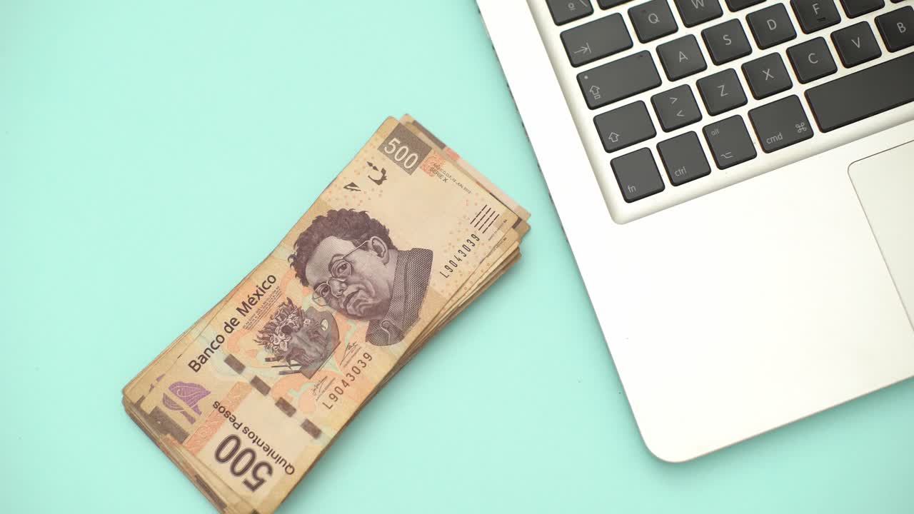 una mano pone el dólar mexicano sobre un escritorio con una computadora portátil - toma estática de alto ángulo de arriba hacia abajo