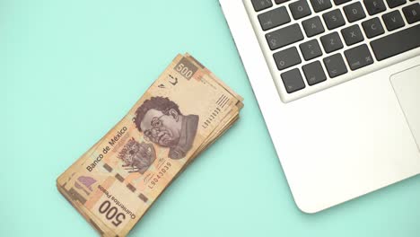 Una-Mano-Pone-El-Dólar-Mexicano-Sobre-Un-Escritorio-Con-Una-Computadora-Portátil---Toma-Estática-De-Alto-ángulo-De-Arriba-Hacia-Abajo