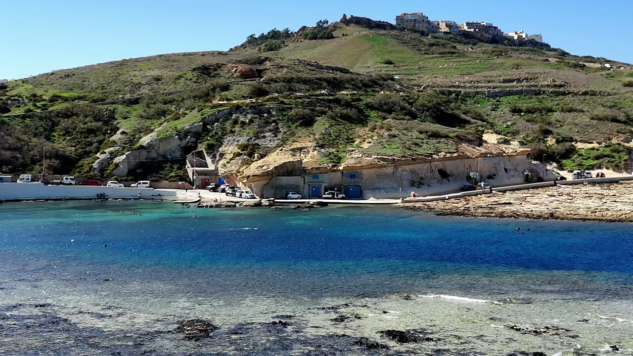 Qbajjar Bay, Gozo, med traditionelle saltpander og båd-huler, der er udhugget i kalkstensklipper under en klar, blå himmel med levende turkisblåt middelhavsvand set fra oven på en fredfyldt sommermorgen