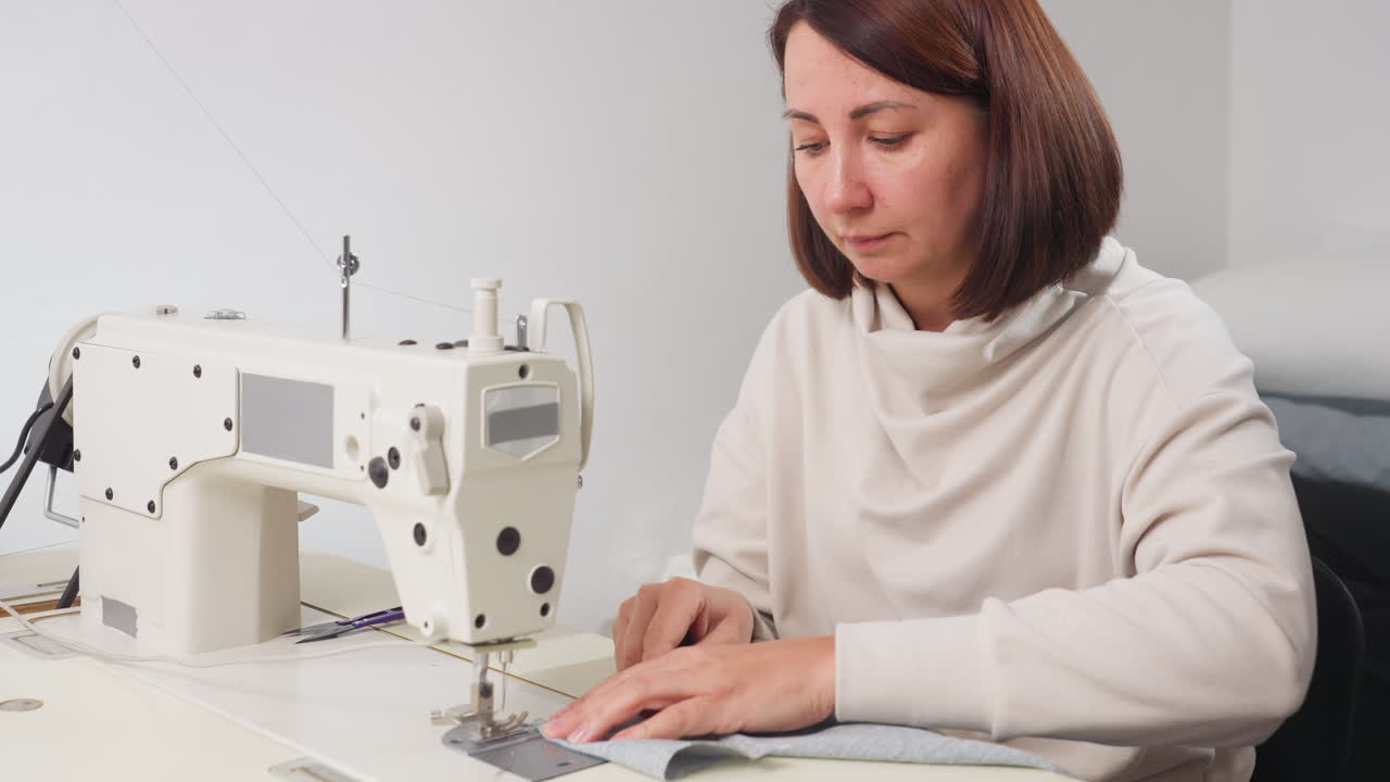 Focused woman in light clothing prepares fabric on white sewing machine in brightly lit workspace, showing precision and attention to detail during beginning of tailoring process