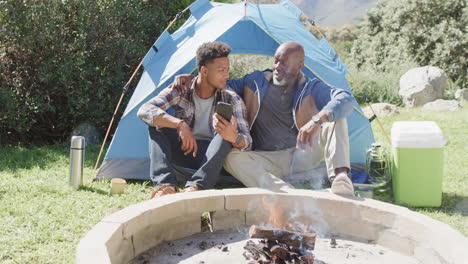 Feliz-Padre-Afroamericano-E-Hijo-Adulto-Sentados-Afuera-Usando-Un-Teléfono-Inteligente-Al-Sol,-En-Cámara-Lenta