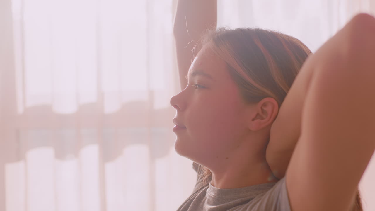 Woman stretches shoulder backward while gently touching hair with closed eyes in warm morning light, creating moment of calm relaxation and physical self care in quiet indoor home environment