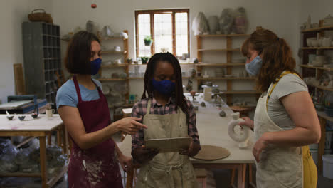 diversas mujeres alfareras con máscara facial y delantal usando tableta digital en el estudio de cerámica