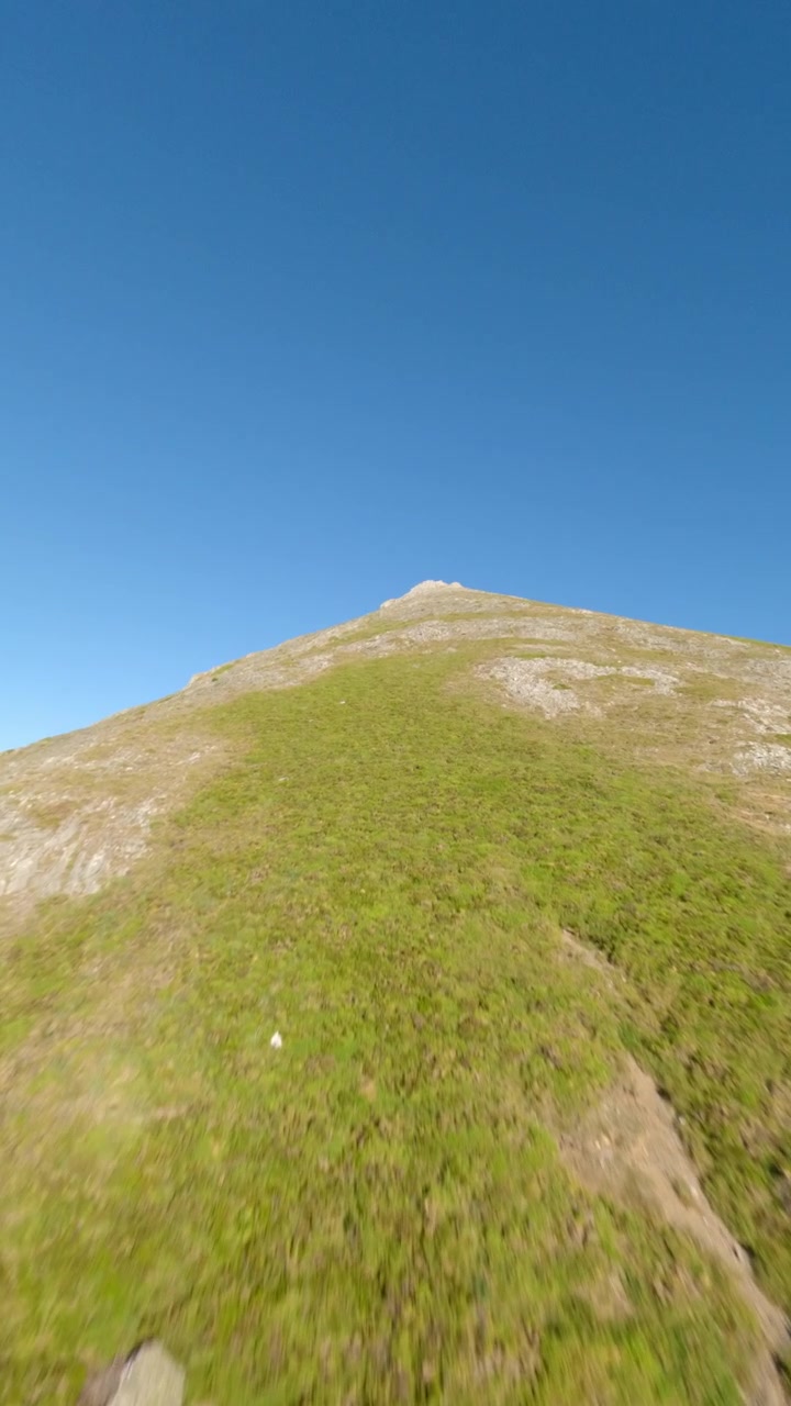 Dinamica ripresa con drone FPV che ascende una cima montuosa rocciosa ed erbosa sotto un cielo azzurro limpido. Ideale per contenuti di avventura all'aperto, benessere e viaggi