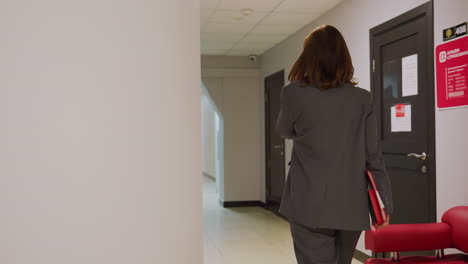 Businesswoman walking down modern office corridor, holding red folder, gray suit, professional attire, office environment, confident career woman, red bench, corporate workspace, stylish business