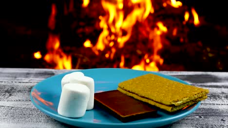 this is a video of a fire burning in the background and a plate with the ingredients to make smores