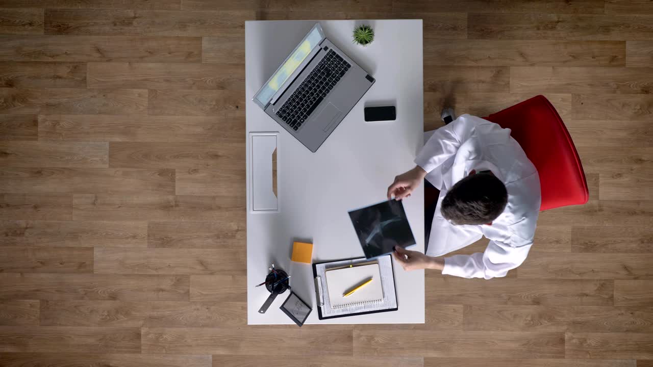 Young doctor in white coat analyzing x-ray scan and typing on laptop, topshot, medical concept