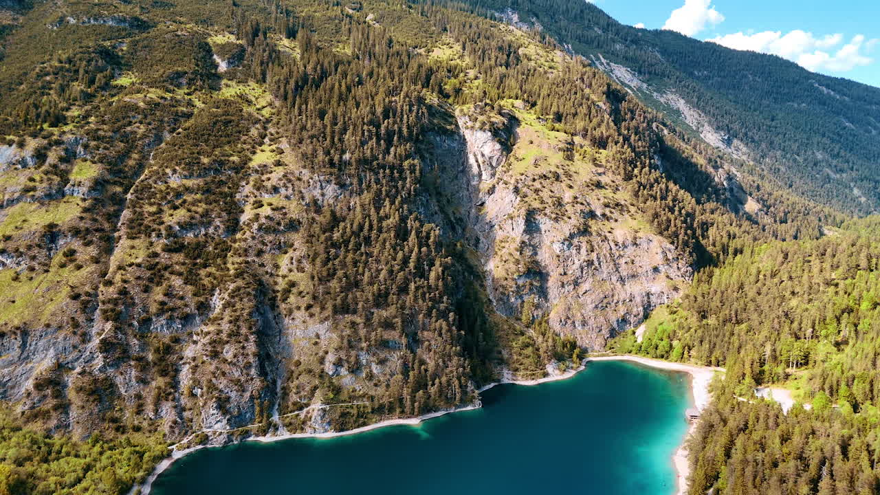 Flying above the turquoise lake in the mountains. View on the unapproachable rocks overgrown with evergreen woods.