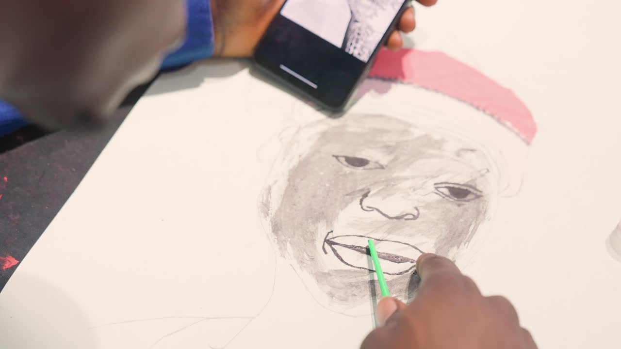 Seniman berkulit hitam membuat sketsa potret dengan referensi ponsel. Membuat studi tonal lukisan hitam di atas kertas, tangan memandu pensil dan arang menambahkan bayangan pada wajah dan topi merah, suasana studio dekat dengan konsentrasi penuh.