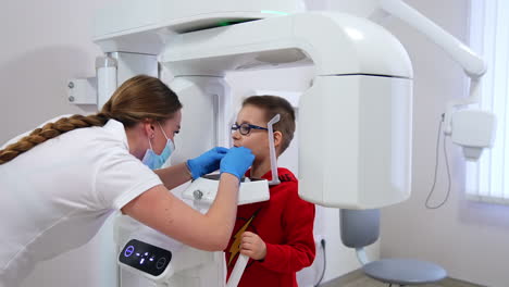 medical worker at dental cabinet preparing little patient to a procedure at x-ray machine. doctor is setting equipment in a mouth of a boy in glasses.