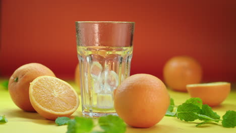 dropping ice blocks in an empty highball glass, surrounded by fresh oranges and spearmint, isolated on a yellow and orange background, slow motion product video