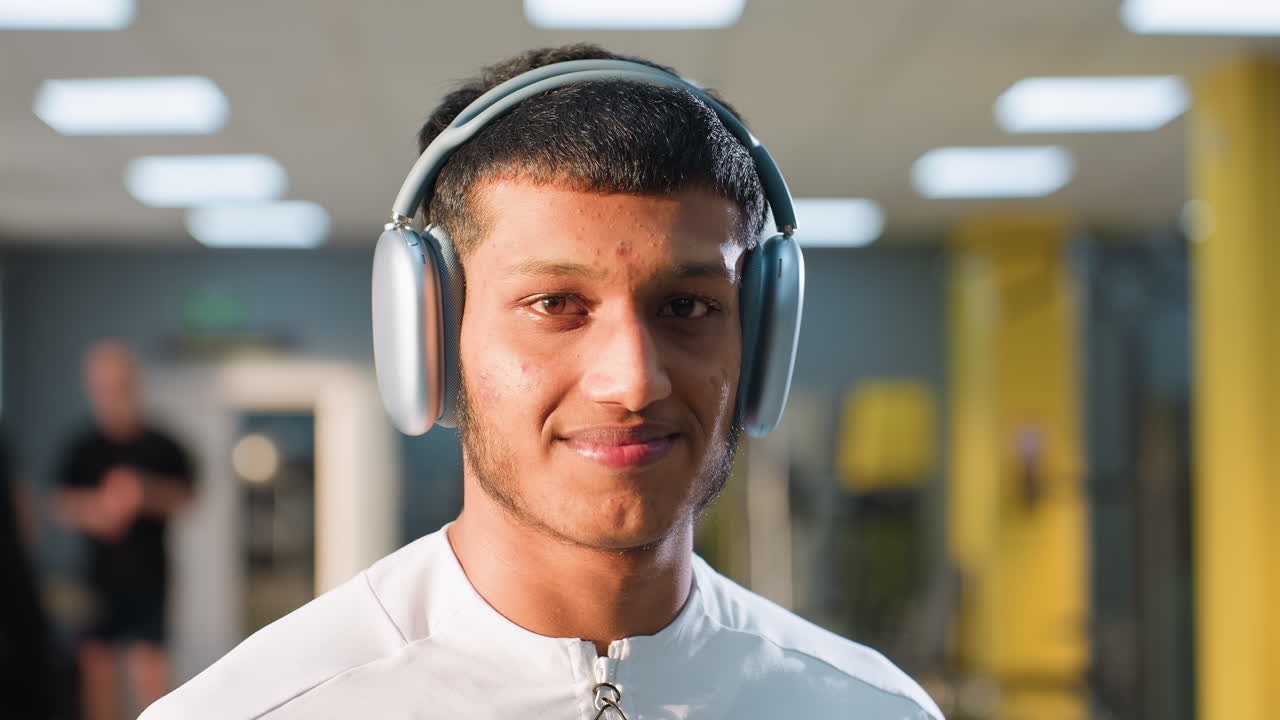 Man wearing white activewear and large headphones poses with elegant and composed look in modern gym setting, soft lighting highlighting facial expression as blurred figures exercise in background