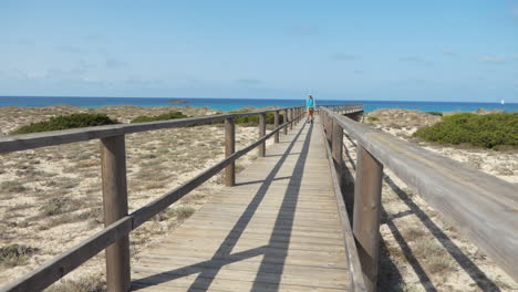 Man-European-tourist-coming-from-the-sea-with-a-blue-towel-on-his-shoulders-by-walking-on-a-path-in-a-wooden-slat,-view-in-perspective