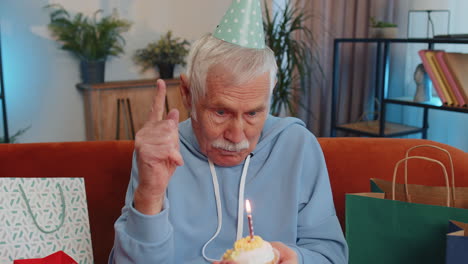 happy elderly old man celebrating birthday party, makes wish blowing burning candle on small cupcake