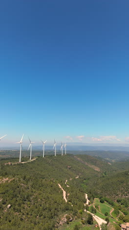 aerial reverse flight showing multiple wind turbines aligned over wooded hills and farmland, with rural paths and a traditional house under a clear blue sky. renewable energy in scenic setting