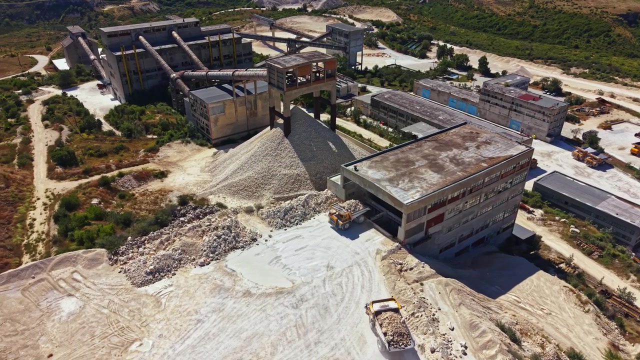 Large construction site shows machinery and materials in production area with nearby structures and landscaping visible in the background