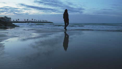 Twilight-beach-with-silhouette-of-man-jumping-over-puddle,-low-tide-reflection